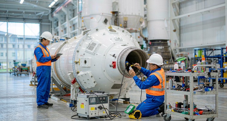 Technicians inspecting and cleaning the engine section of a recovered rocket booster on a horizontal stand, with NDT equipment and a tool cart
